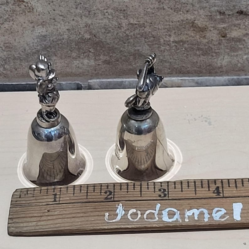 Pu&ograve; includere: Due campanelli color argento con figurine di animali sulla parte superiore. I campanelli sono appoggiati su basi di plastica trasparente. Un righello di legno con la scritta "Jodamel" &egrave; in primo piano. I campanelli sono alti circa 7.6 cm.