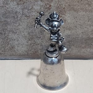Könnte beinhalten: Eine silberfarbene Glocke mit einer Mickey Mouse-Figur in einer Marschkapellenuniform. Die Figur hält einen Taktstock und trägt einen Hut. Die Glocke hat eine klassische Form und eine polierte Oberfläche, ideal für Sammler.