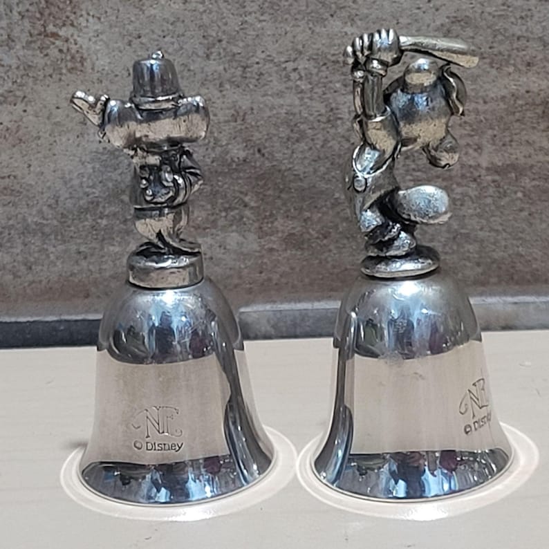 Pu&ograve; includere: Due campane color argento con figurine di personaggi Disney. Una campana presenta un personaggio con cappello e braccio teso, l'altra un personaggio che fa oscillare una mazza. Ogni campana ha il logo Disney e un "NC" stilizzato.