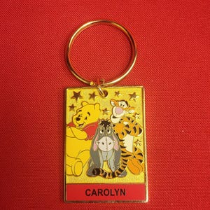 May include: A gold-colored rectangular keychain featuring Winnie the Pooh, Tigger, and Eeyore. The characters are set against a yellow background with star accents. The name "CAROLYN" is printed in red at the bottom. A gold ring is attached at the top.