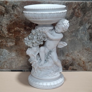 Könnte beinhalten: Weiße Cherub-Statue mit großem schalenförmigem Oberteil. Der Cherub hält ein Blumengesteck und sitzt auf einem felsigen Sockel. Die Statue hat ein detailliertes, klassisches Design und eignet sich für die Innen- oder Außendekoration.