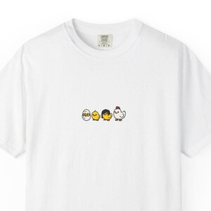 May include: White t-shirt with a small graphic of three cartoon chicks and an egg. The chicks are yellow, black, and white, with one hatching from an egg. The shirt has a crew neck and short sleeves. The brand name "Comfort Colors" is printed on the upper back.