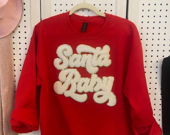 May include: A bright red sweatshirt with the words "Santa Baby" in a white, textured font with gold accents. The sweatshirt is hanging on a hanger against a white background.