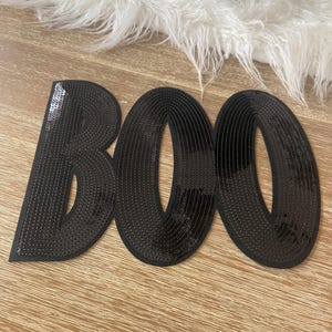 Può includere: Lettere "BOO" nere con paillettes su una superficie di legno. Le lettere sono cucite con filo nero e hanno un aspetto lucido. Le lettere sono disposte orizzontalmente, perfette per le decorazioni di Halloween.