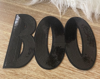 May include: Black sequined "BOO" letters on a wooden surface. The letters are stitched with black thread and have a shiny appearance. The letters are arranged horizontally, perfect for Halloween decor.