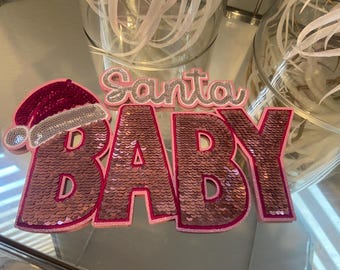 May include: Pink and white sequined Christmas decoration with the text "Santa Baby".