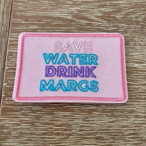 May include: A rectangular pink patch with the embroidered text "SAVE WATER DRINK MARGS" in turquoise, purple, and pink. The patch has a pink border and is likely designed to be sewn or ironed onto clothing or accessories.