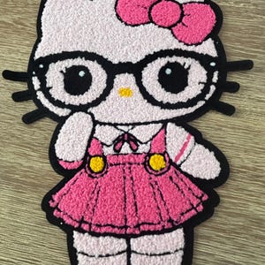 Peut inclure: Tapis rose et blanc représentant un personnage de chat de dessin animé portant des lunettes et un nœud rose. Le chat porte une robe rose avec des boutons jaunes et un contour noir. Le tapis est sur une surface en bois.
