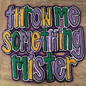 May include: A colorful patch with the phrase "THROW ME SOMETHING MISTER" in a playful, embroidered style. The text features a combination of purple, green, yellow, and white, with a black outline. The patch is likely for clothing or accessories.