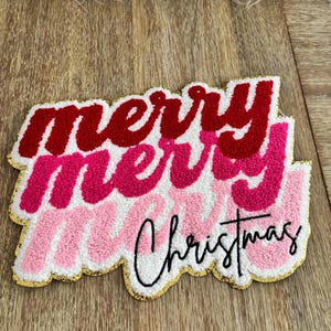 Puede incluir: Adorno navideño con la frase "merry merry Christmas" en rojo y rosa superpuestos, con borde dorado. El texto está sobre un fondo blanco con una textura en relieve.