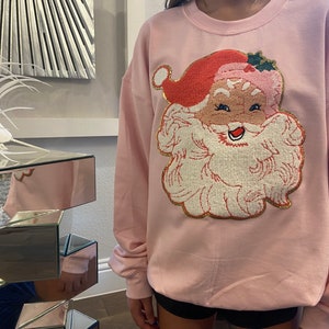 Puede incluir: Sudadera rosa claro con un diseño bordado de Papá Noel. Papá Noel tiene un gorro rojo, barba blanca y una cara sonriente. La sudadera tiene mangas largas y cuello redondo, perfecta para la temporada navideña.