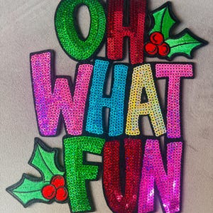 May include: A sequined patch featuring the text OH WHAT FUN in multicolored letters including green, red, blue, yellow, and pink. The design is accented with two sprigs of green holly leaves and red berries, all outlined in black thread.