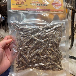 Puede incluir: Una bolsa de plástico transparente llena de anchoas medianas secas. La bolsa tiene una etiqueta amarilla y naranja con el texto "MEDIUM DILIS DRIED MEDIUM ANCHOVIES 100g" y "TUYO DE CAPIZ". La etiqueta también indica "FROM THE SEAFOOD CAPITAL ROXAS CITY CAPIZ, PHILIPPINES".