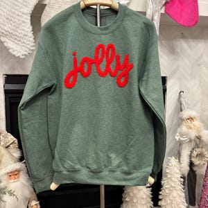 May include: A green crewneck sweatshirt with the word "jolly" in red, textured lettering. The sweatshirt is displayed on a hanger. The background includes Christmas stockings and decorations, suggesting a holiday theme.