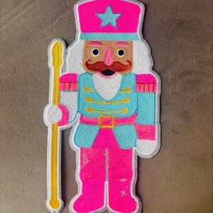 Könnte beinhalten: Ein mit Pailletten besetzter Nussknacker-Aufnäher mit einem leuchtend pinken Hut, einem hellblauen Stern, einer hellblauen Jacke mit Goldverzierungen und pinker Hose. Die Figur hat ein beiges Gesicht, einen roten Schnurrbart und hält einen goldenen Stab.