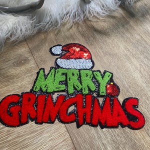 Puede incluir: Decoración navideña con las palabras "MERRY GRINCHMAS" en letras rojas y verdes brillantes. El diseño incluye un gorro de Papá Noel y un pequeño adorno, perfecto para la decoración navideña.