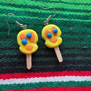 May include: A pair of yellow popsicle-shaped earrings with blue and orange details. The earrings are made of polymer clay and have silver hooks.