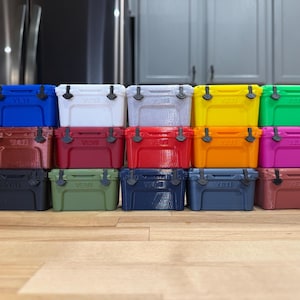 May include: A collection of small, colorful coolers in various shades, including blue, white, gray, yellow, green, red, orange, and pink. Each cooler has a black handle and latch. The coolers are arranged in rows on a wooden surface.