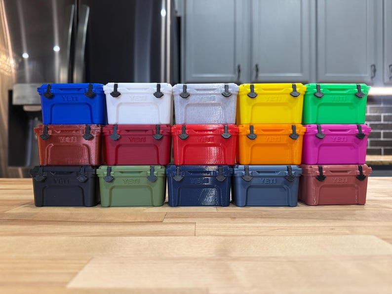 May include: A collection of small, colorful coolers in various shades, including blue, white, gray, yellow, green, red, orange, and pink. Each cooler has a black handle and latch. The coolers are arranged in rows on a wooden surface.