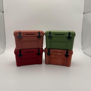 May include: Four small, colorful coolers stacked together. The coolers are in shades of red, orange, and green, with black latches and the brand name "YETI" printed on the front. The coolers are on a white surface.