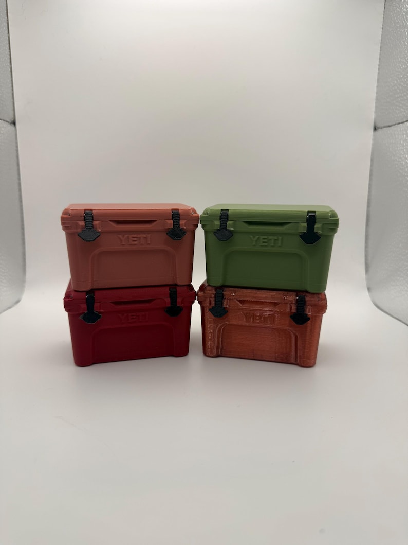 May include: Four small, colorful coolers stacked together. The coolers are in shades of red, orange, and green, with black latches and the brand name "YETI" printed on the front. The coolers are on a white surface.