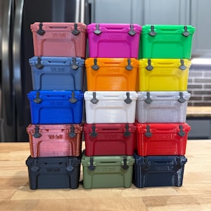 May include: A stack of colorful, miniature coolers in various shades including pink, green, blue, and red. Each cooler has a black handle and the word "Yeti" printed on the front. The coolers are arranged on a wooden surface.