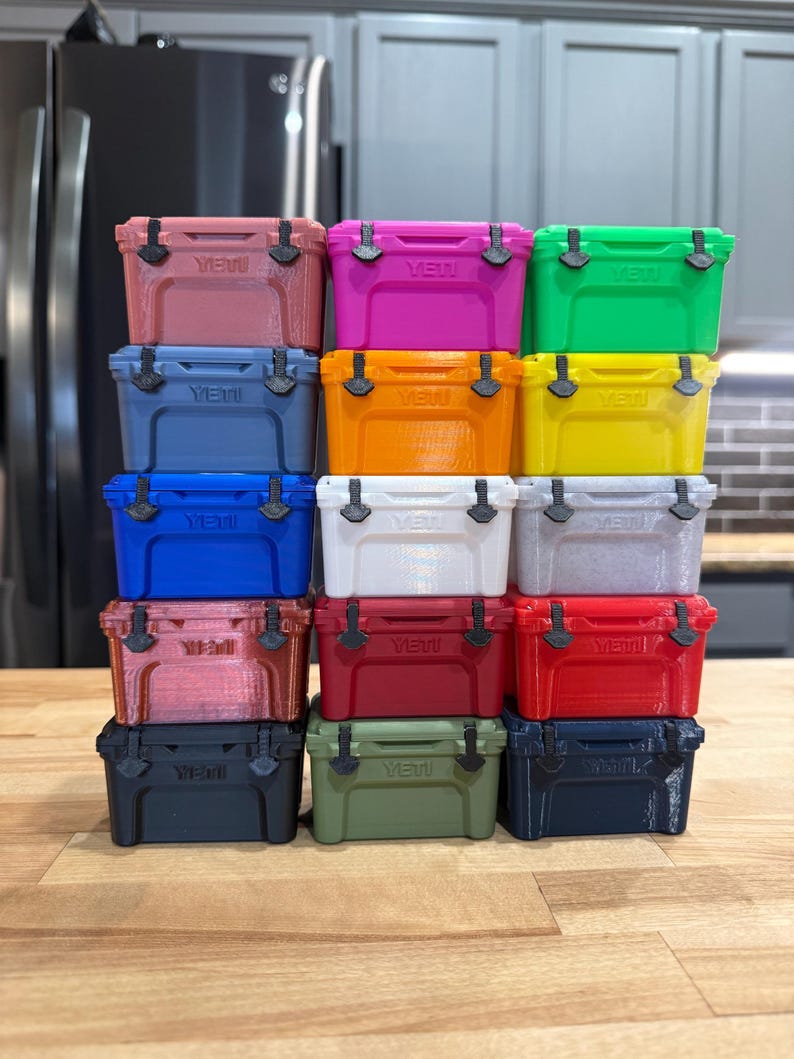 May include: A stack of colorful, miniature coolers in various shades including pink, green, blue, and red. Each cooler has a black handle and the word "Yeti" printed on the front. The coolers are arranged on a wooden surface.