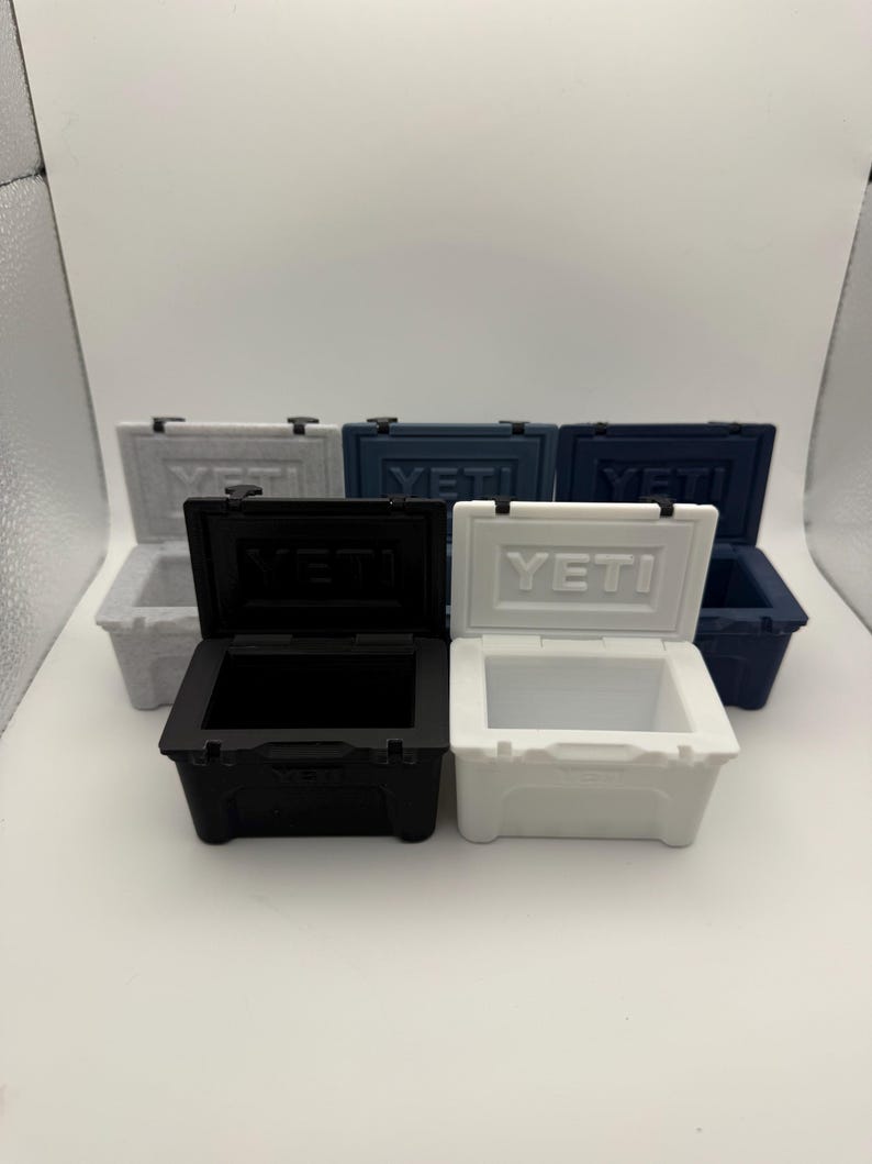 May include: A collection of small, hard-sided coolers in various colors, including white, black, and blue. Each cooler has a hinged lid and the brand name "YETI" is visible on the front. The coolers are designed to keep items cold.