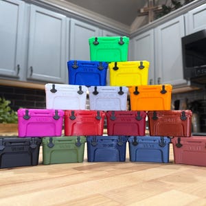 May include: A collection of colorful, small coolers in various shades, including green, blue, yellow, white, pink, red, and brown. Each cooler has a black handle and latch. The coolers are stacked on a wooden surface.