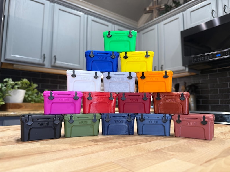 May include: A collection of colorful, small coolers in various shades, including green, blue, yellow, white, pink, red, and brown. Each cooler has a black handle and latch. The coolers are stacked on a wooden surface.