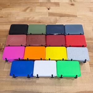 May include: A collection of rectangular, colorful cases with black hinges. The cases are arranged in rows and columns, showcasing a variety of colors including black, green, blue, red, pink, orange, yellow, and gray. The cases are on a wooden surface.