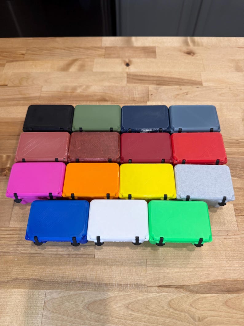 May include: A collection of rectangular, colorful cases with black hinges. The cases are arranged in rows and columns, showcasing a variety of colors including black, green, blue, red, pink, orange, yellow, and gray. The cases are on a wooden surface.