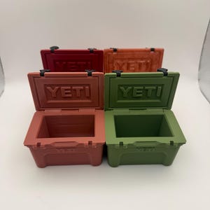 May include: Four small, open-top coolers in shades of red, orange, and green. Each cooler has the word "YETI" embossed on the lid. The coolers are arranged on a white surface, with the lids of the front two coolers open.