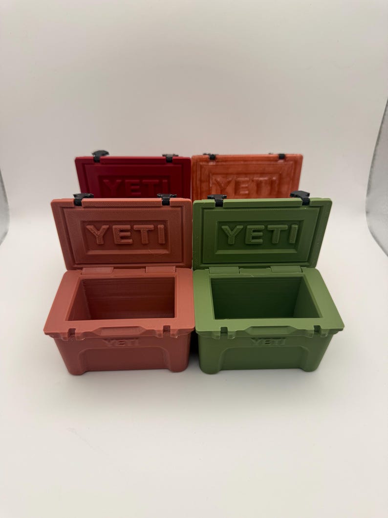 May include: Four small, open-top coolers in shades of red, orange, and green. Each cooler has the word "YETI" embossed on the lid. The coolers are arranged on a white surface, with the lids of the front two coolers open.