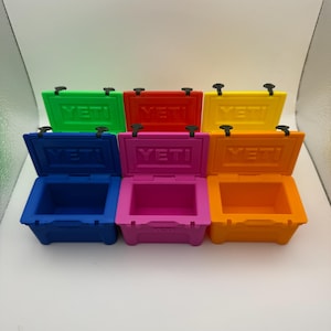 May include: A collection of small, colorful coolers in green, red, yellow, blue, pink, and orange. Each cooler has a hinged lid and the brand name "YETI" printed on the lid. The coolers are arranged in two rows on a white surface.