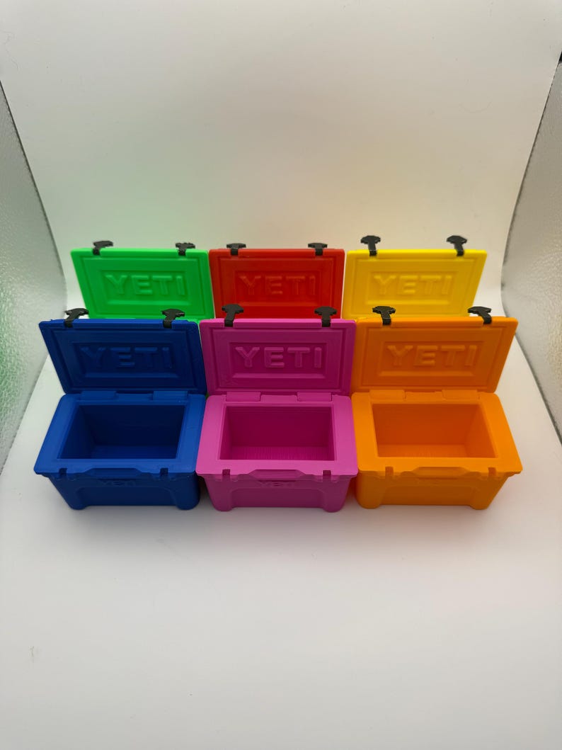 May include: A collection of small, colorful coolers in green, red, yellow, blue, pink, and orange. Each cooler has a hinged lid and the brand name "YETI" printed on the lid. The coolers are arranged in two rows on a white surface.