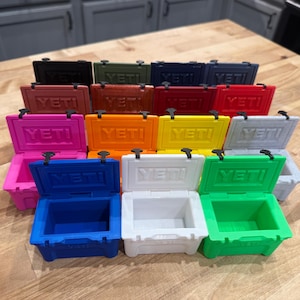 May include: A collection of small, colorful coolers in various shades, including black, green, blue, pink, and orange. Each cooler has the word "YETI" printed on the front. The coolers are arranged on a wooden surface.