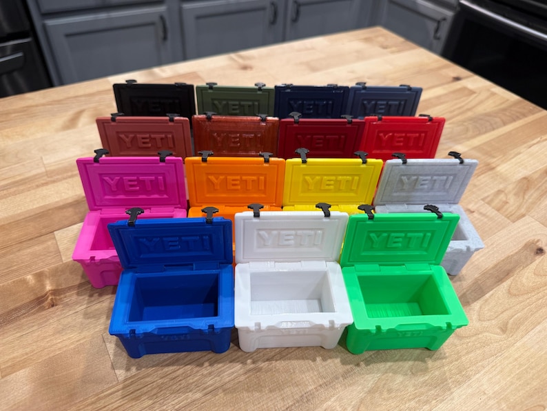 May include: A collection of small, colorful coolers in various shades, including black, green, blue, pink, and orange. Each cooler has the word "YETI" printed on the front. The coolers are arranged on a wooden surface.