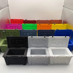 May include: A collection of colorful, small, insulated coolers. The coolers are in various colors, including red, pink, green, blue, black, and white. Each cooler has the brand name "YETI" on the front.