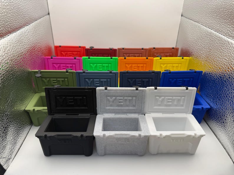 May include: A collection of colorful, small, insulated coolers. The coolers are in various colors, including red, pink, green, blue, black, and white. Each cooler has the brand name "YETI" on the front.