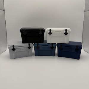 May include: A collection of small, hard-sided coolers in various colors: black, white, gray, navy blue, and a darker navy blue. Each cooler has a black handle and latch. The brand name "YETI" is visible on the front of each cooler.