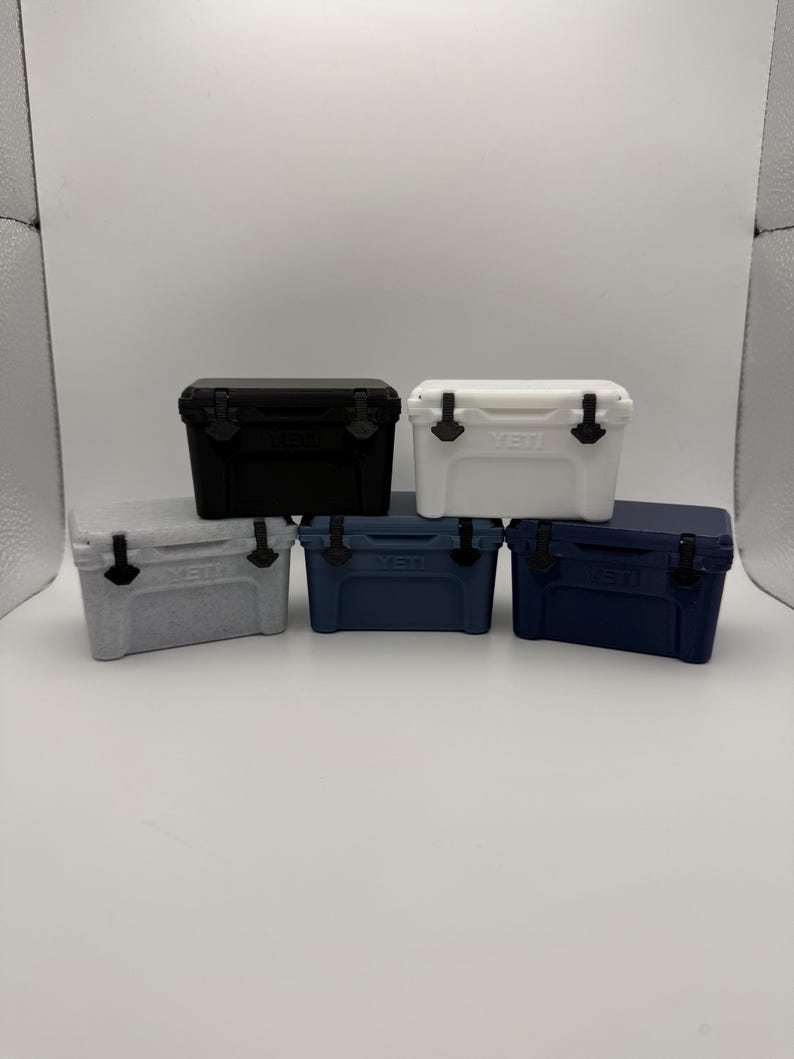 May include: A collection of small, hard-sided coolers in various colors: black, white, gray, navy blue, and a darker navy blue. Each cooler has a black handle and latch. The brand name "YETI" is visible on the front of each cooler.