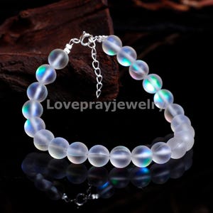 Puede incluir: Un brazalete con cuentas redondas, transparentes e iridiscentes. Las cuentas tienen un sutil brillo de arcoíris. El brazalete está ensartado en una cadena plateada con un cierre ajustable. El texto "Loveprayjewel" es visible.