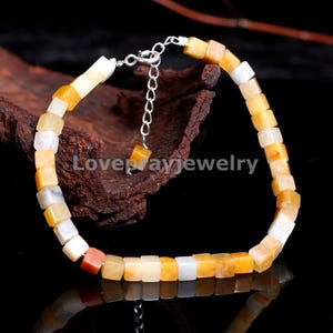 Puede incluir: Un brazalete de cuentas cuadradas de color amarillo, blanco y rojo con un cierre plateado. El brazalete está hecho de cuentas de piedra natural y tiene una longitud aproximada de 18 cm.