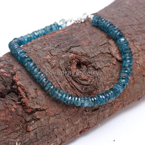 Puede incluir: Una pulsera de piedras preciosas azules con cierre plateado. La pulsera está hecha de pequeñas cuentas facetadas y se exhibe sobre un trozo de madera marrón oscuro. Las cuentas son de un color verde azulado intenso.