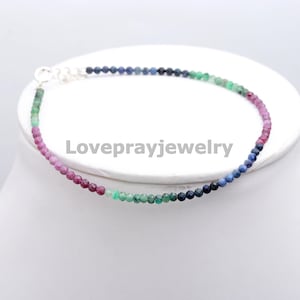 Puede incluir: Una delicada pulsera compuesta por pequeñas cuentas de colores. Las cuentas cambian de color, pasando del rosa al verde, luego al azul y finalmente a un azul más oscuro. La pulsera está ensartada en una fina cadena de plata y se muestra sobre una superficie blanca.