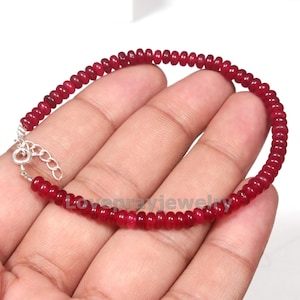 May include: A red beaded bracelet with a silver clasp. The bracelet is made of small, round, red beads strung together. The clasp is a simple hook and eye design.