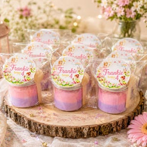 May include: A set of small clear jars filled with layered pink and purple powder, each topped with a round sticker featuring a fairy illustration and the text Frankie turns 4, accompanied by a small wooden spoon, all individually wrapped in plastic.