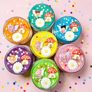 May include: Seven round containers of slime in various colours, each decorated with a fairy, mushroom house, and daisy. The slime colours are orange, green, blue, yellow, purple, pink, and green. The containers are arranged on a pink surface.