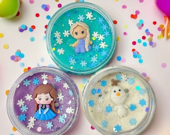 May include: Three round clear containers filled with colorful slime. One container has blue slime with a blonde character charm and white snowflake glitter. Another has purple slime with a brown-haired character charm and snowflake glitter. The third has white slime with a snowman character charm and blue snowflake glitter.